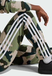 Pantaloni della tuta in stampa camouflage verde e beige con strisce bianche laterali; tessuto morbido, vestibilità rilassata, abbinati a una sneakers nera.