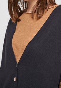 Marinblå v-ringad cardigan med en texturerad finish, som har en brun topp under. Två knappar pryder cardiganens framsida.