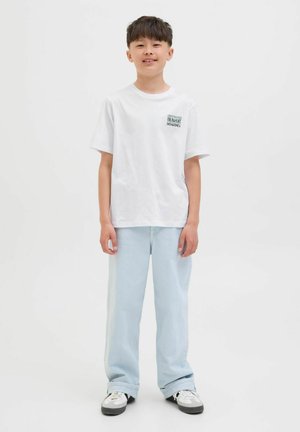 Weißes Baumwoll-T-Shirt mit einem kleinen Motiv auf der linken Brust, kombiniert mit hellblauen, baggy Jeans und schwarz-weißen Sneakers.