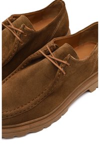 Brown suede shoes with a smooth texture, rounded shape, and beige rubber soles; features laces and contrasting stitching details.