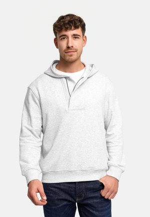 Jeune homme aux cheveux courts et bouclés portant un sweat à capuche gris clair et un jean foncé, debout devant un fond blanc uni.