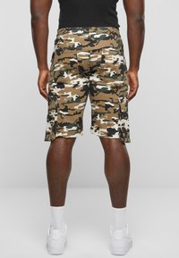 Camo kravas šorti brūnā, zaļā un krēmkrāsā. Iespējas sānu kabatas un elastīgs josta. Valkāti ar baltām sporta apaviem un īsām zeķēm.