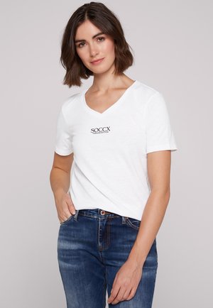 Weißes V-Ausschnitt-T-Shirt mit kurzen Ärmeln und dem schwarzen Logo "SOCX TIME-LESS EDITION". Kombiniert mit blauen Denim-Jeans.
