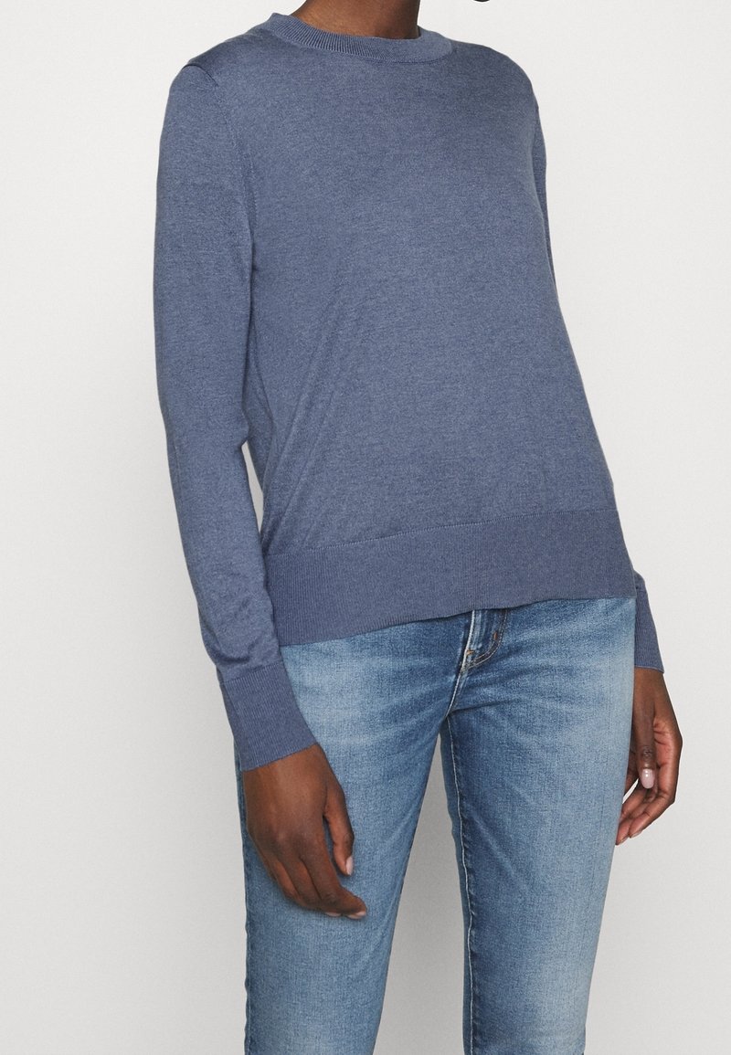 Personne portant un pull à col rond à manches longues bleu et un jean en denim bleu clair, debout devant un fond blanc uni.