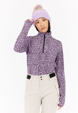 Langsleeve, hoge kraag, paarse luipaardprint top met zwarte rits; gecombineerd met beige broek met zwarte accenten. Warme gebreide lavendelkleurige muts.