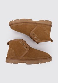 Botas de tobillo de ante marrón con cordones, suelas de goma y detalles de costura, exhibidas sobre un fondo claro y liso.