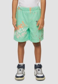 Mintgrüne Shorts mit orange-weißer Spritzmuster, elastischem Bund, lockerer Passform und seitlichen Taschen. Kombiniert mit hohen Turnschuhen.