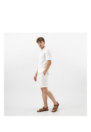 Homme debout regardant sur le côté, portant une chemise blanche à manches courtes, un short blanc et des sandales marron sur un fond clair uni.