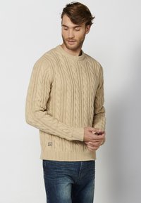 Maglione beige a maglia con un motivo testurizzato, collo rotondo e polsini a coste, abbinato a jeans in denim blu. Piccolo logo sul lato inferiore.