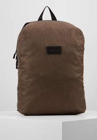 Zaino in stoffa marrone con una finitura testurizzata, forma rettangolare, maniglia superiore e una patch in pelle nera con logo sul davanti.