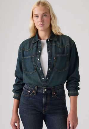 Junge Frau mit blondem Haar trägt eine dunkle Jeansjacke über einem weißen Hemd und dunkle Jeans, steht vor einem schlichten hellen Hintergrund.
