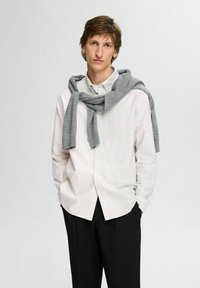 Camicia bianca a maniche lunghe con leggere strisce pastello, colletto abbottonato, abbinata a pantaloni scuri e un maglione grigio appoggiato sulle spalle.