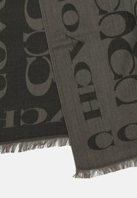 Harmaa kudottu huivi, jossa on suuria mustia "C"-logokuvioita, viimeistelty risareunoilla ja tekstuuripinnalla.