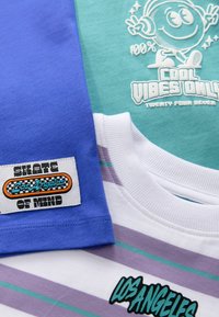 Tres camisetas: una azul, una verde azulado con un gráfico blanco y una blanca con rayas moradas y un parche de logo, que exhiben diferentes diseños y texturas.