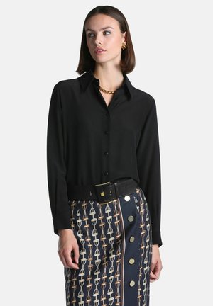 Femme portant une chemise noire boutonnée avec des boucles d'oreilles et un collier dorés, une jupe à motifs avec de gros boutons et une large ceinture noire.