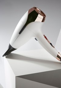 Sèche-cheveux blanc avec une buse noire, des accents rose gold et des boutons de contrôle. Design épuré avec une finition brillante. Monté sur une surface géométrique.