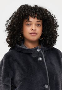 Veste à capuche en fausse fourrure de couleur gris foncé. Présente de grands boutons, une texture douce et une coupe décontractée. Accent sur la chaleur et le confort.