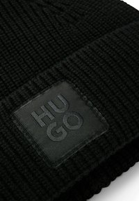 Berretto lavorato a maglia nero con texture a coste, dotato di una prominente etichetta quadrata in pelle nera impressa con la scritta "HUGO" sul davanti.