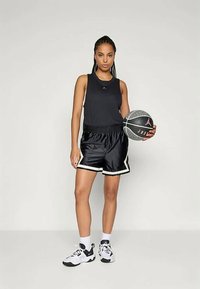 Čierny športový tank top a lesklé čierne basketbalové šortky s bielymi akcentmi; drží textúrovanú basketbalovú loptu s ružovým logom; biele športové topánky.