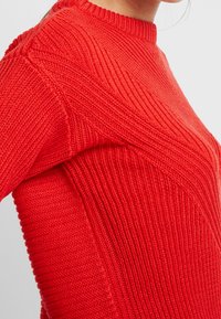 Czerwony sweter z dzianiny z fakturowanym wzorem, okrągłym dekoltem oraz dopasowanym krojem z prążkowanymi detalami na ramionach.