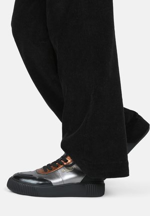 Sorte corduroybukser med en metallisk sølv- og orange sneaker, der har en tekstureret overflade, sorte snørebånd og en tykk gummisål.