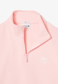 Pullover a mezze zip di colore rosa chiaro realizzato in morbido tessuto. Presenta un logo bianco Lacoste sul petto e un colletto interno testurizzato. Vestibilità unisex.