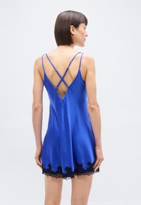 Γυναίκα που φοράει μπλε σατέν φόρεμα με λεπτή μαύρη δαντέλα, με σταυρωτές λεπτές τιράντες και βαθύ V στο πίσω μέρος.