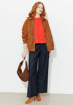 Femme aux cheveux roux portant une veste en daim marron, un haut rouge, un pantalon large bleu marine, des bottines à talons en daim marron, et tenant un sac à main imprimé léopard.