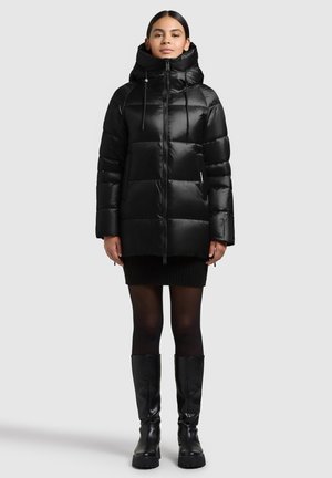 Frau steht und trägt eine schwarze Steppjacke mit hohem Kragen und Kapuze, einen schwarzen Minirock, schwarze Strumpfhosen und kniehohe schwarze Stiefel.