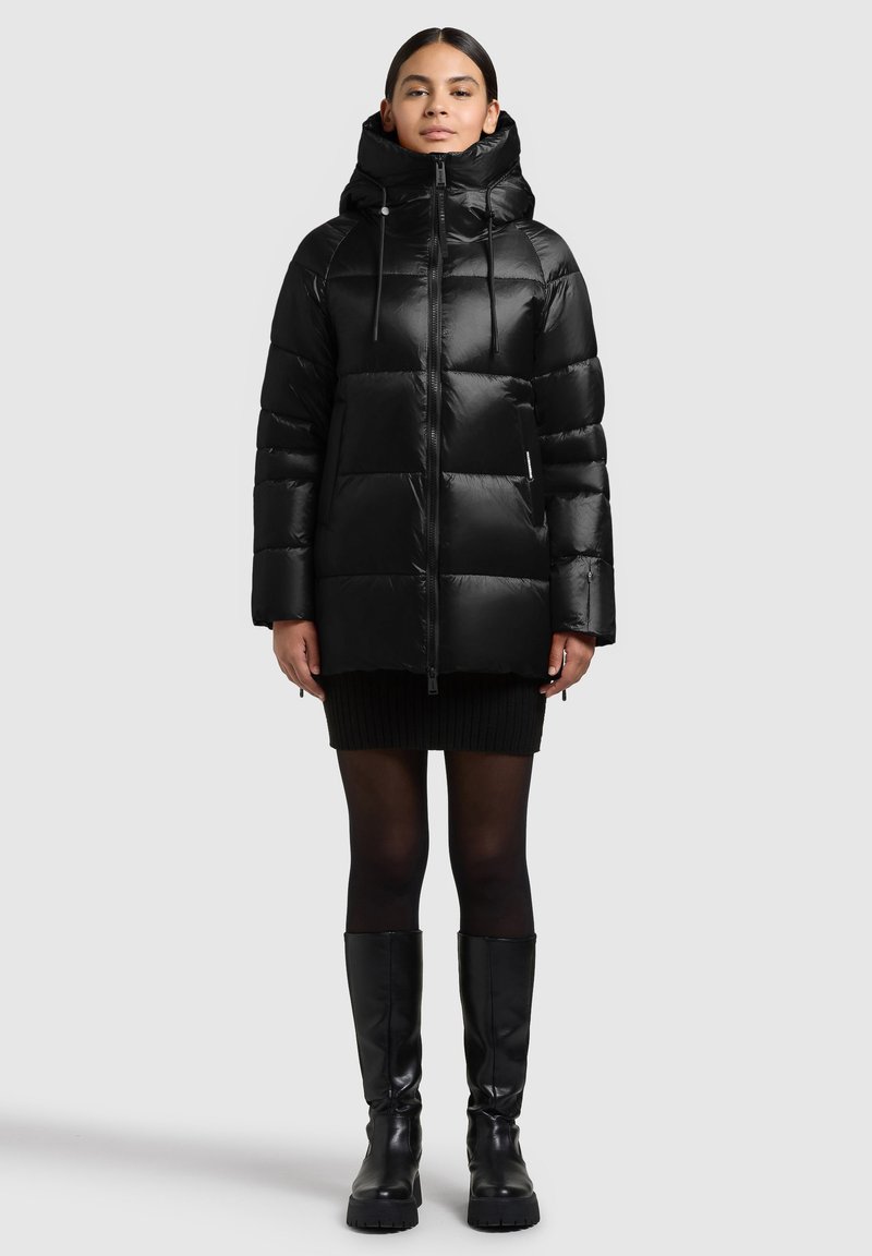 Kobieta stojąca, ubrana w czarną pikowaną kurtkę z wysokim kołnierzem i kapturem, czarną mini spódniczkę, czarne rajstopy oraz czarne kozaki za kolano.