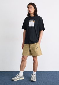 Svart kortermet t-skjorte med grafisk trykk, kombinert med khaki-shorts med brodert logo. Modellen har på seg grå sneakers og hvite sokker.