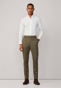 Witte overhemd met knopen, slim-fit olijfgroene broek en bruine enkellaarsjes. De stof lijkt glad; de broek heeft voorzakken en een ritssluiting.