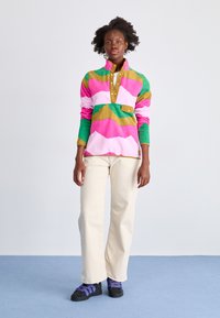 Maglione in pile colorato con motivi a onde rosa, verdi e dorati, indossato sopra una camicia button-up marrone chiaro. Abbinato a pantaloni crema e scarpe nere.