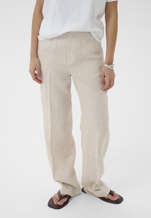 Personne portant un pantalon ample en lin beige, une chemise blanche à manches courtes, des tongs noires à bout carré et un bracelet en argent, debout sur un fond blanc.