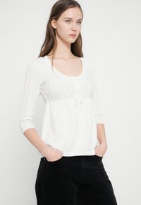 Λευκή πλεκτή μπλούζα με στρογγυλή λαιμόκοψη, κουμπωτή μπροστινή πλευρά και ρυθμιζόμενο κορδόνι στη μέση. Διαθέτει μανίκια τριών τετάρτων και υφή με ραβδώσεις.