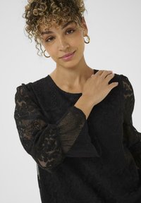 Mlada ženska z curly lasmi, ki nosi zlate uhane na obroč, in črno čipkasto bluzo, nežno dotika svojega ramena ob enobarvni ozadju.