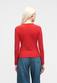 Γυναίκα με μακριά ξανθά μαλλιά, φορώντας εφαρμοστό κόκκινο μπλουζάκι με μακριά μανίκια και μπλε τζιν, από πίσω σε απλό φόντο.