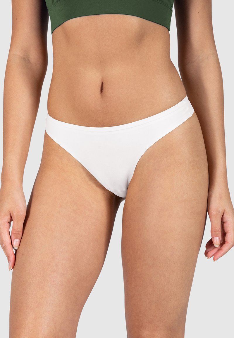 Kobieta w białych majtkach bikini i zielonym krótkim topie, stojąca z luźno opuszczonymi rękami przy bokach na jednolitym tle.