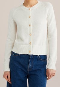 Vit stickad kofta med rund hals och åtta träknappar framtill. Har ribbade manschetter och fåll, mjuk textur och långa ärmar.