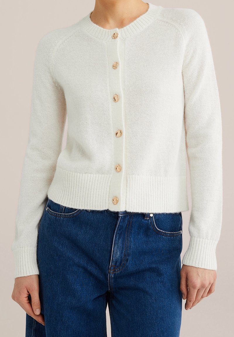Vit stickad kofta med rund hals och åtta träknappar framtill. Har ribbade manschetter och fåll, mjuk textur och långa ärmar.