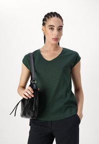 Green short-sleeve v-neck top made of a textured fabric, paired with a black pleated bag hanging from a shoulder strap.
