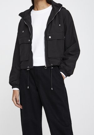 Personne portant une veste à capuche noire raccourcie avec cordons, deux poches avant avec rabat et une chemise blanche en dessous, associée à un pantalon noir.