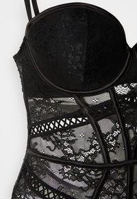 Μαύρο κορμάκι με περίτεχνα σχέδια δαντέλας και τούλι, δομημένα cups και διακοσμητικές τιράντες που σχηματίζουν τη σιλουέτα.