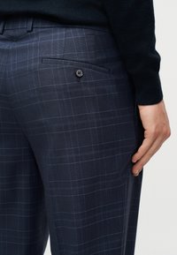 Κοντινό ενός χεριού δίπλα σε σκούρο μπλε παντελόνι με καρό σχέδιο, με κουμπωμένη πίσω τσέπη και μαύρη μπλούζα.