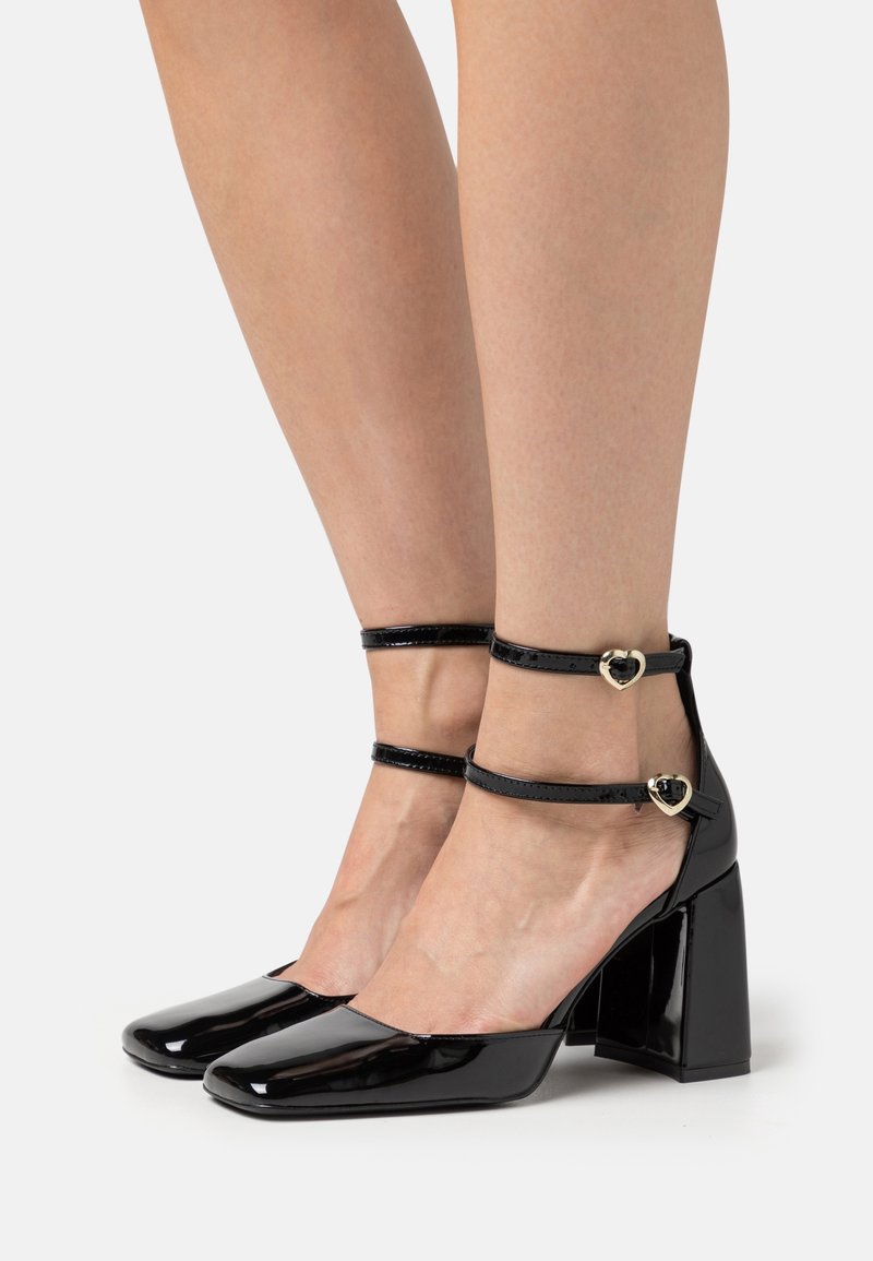 Chaussures à talons blocs en cuir verni noir avec brides à la cheville et boucles dorées en forme de cœur, portées pieds nus.