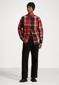 Κόκκινο και μαύρο πουκάμισο καρό με κουμπιά και δύο τσέπες στο στήθος, σε συνδυασμό με μαύρο παντελόνι και μαύρο σκουφάκι, φορεμένα με μαύρα παπούτσια.