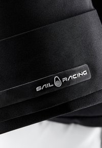 Svart tygjacka med en slät textur som har en rektangulär Sail Racing-logotyp i silver på en svart bakgrund.