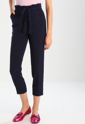 Femme portant un pantalon court bleu marine avec une taille paperbag et des mocassins métalliques roses, debout avec une main dans la poche.