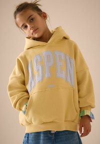 Żółta bluza z kapturem z białym logo "ASPEN" oraz napisem 1982 poniżej. Wykonana z miękkiego materiału, o oversize'owym kroju, z kieszenią kangurką oraz warstwowymi ściągaczami w paski.