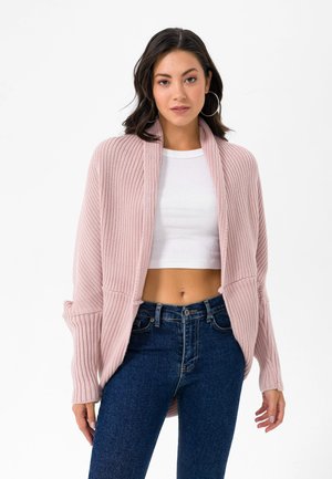 Femme portant de grandes boucles d'oreilles créoles, un cardigan en maille rose clair sur un crop top blanc, et un jean taille haute bleu foncé, debout devant un fond blanc.